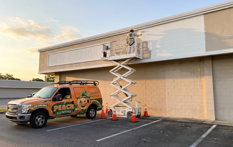Peach Painting - Commercial vs Residential Painting in Tampa: Same Paint, New Rules Painter on a scissor lift painting the block exterior of a commercial building, with a Peach Painting truck and safety cones