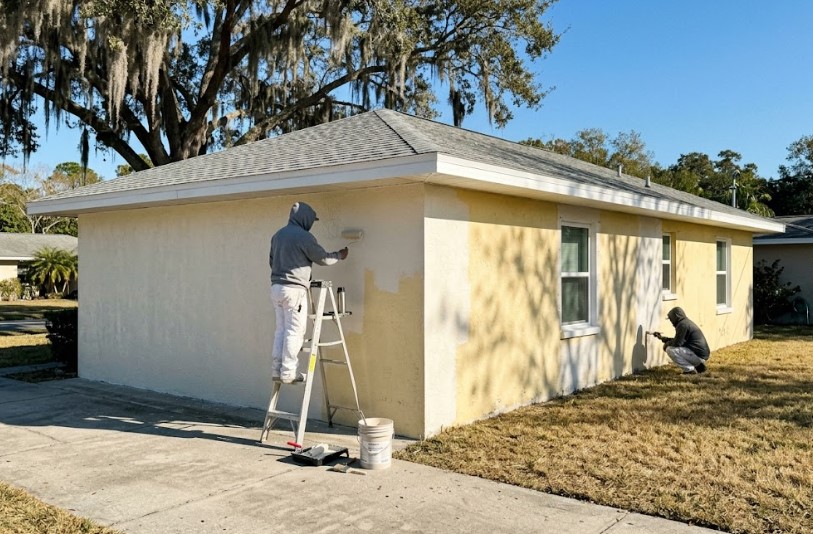 Two painters in hoodies applying cream-colored paint over yellow stucco on a single-story home under a clear blue sky