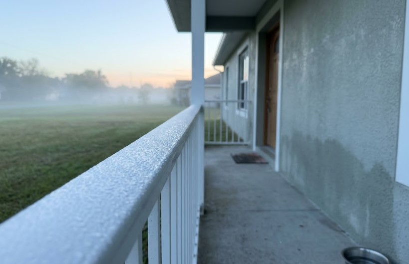 White porch railing covered in beaded moisture at sunrise, with fog over the lawn and a stucco wall beside the porch